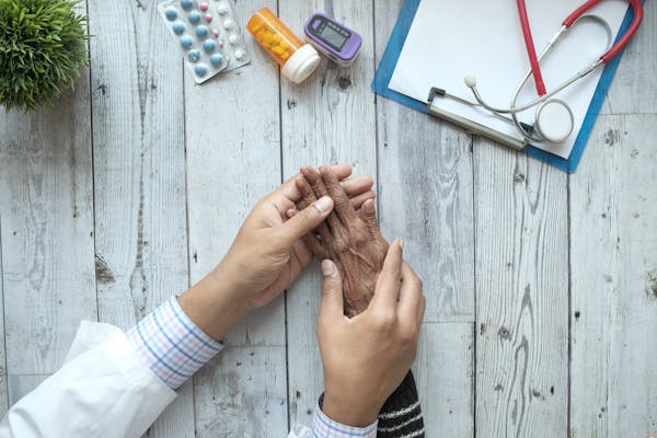 A clinician's hand rests gently on an elderly patient's hand beside a stethoscope