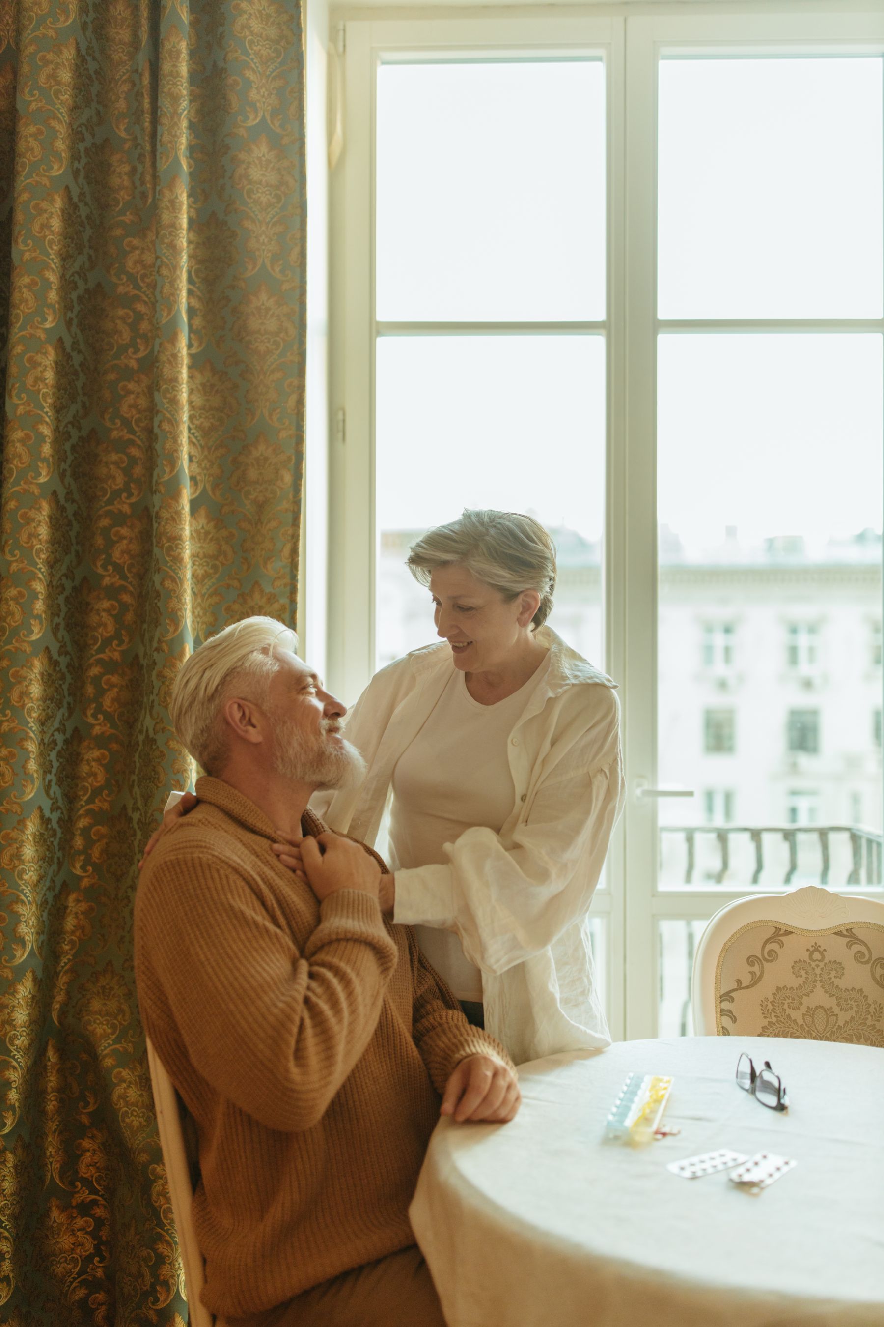 An older couple at home in warm morning light; she rests a hand on his shoulder beside his morning medications